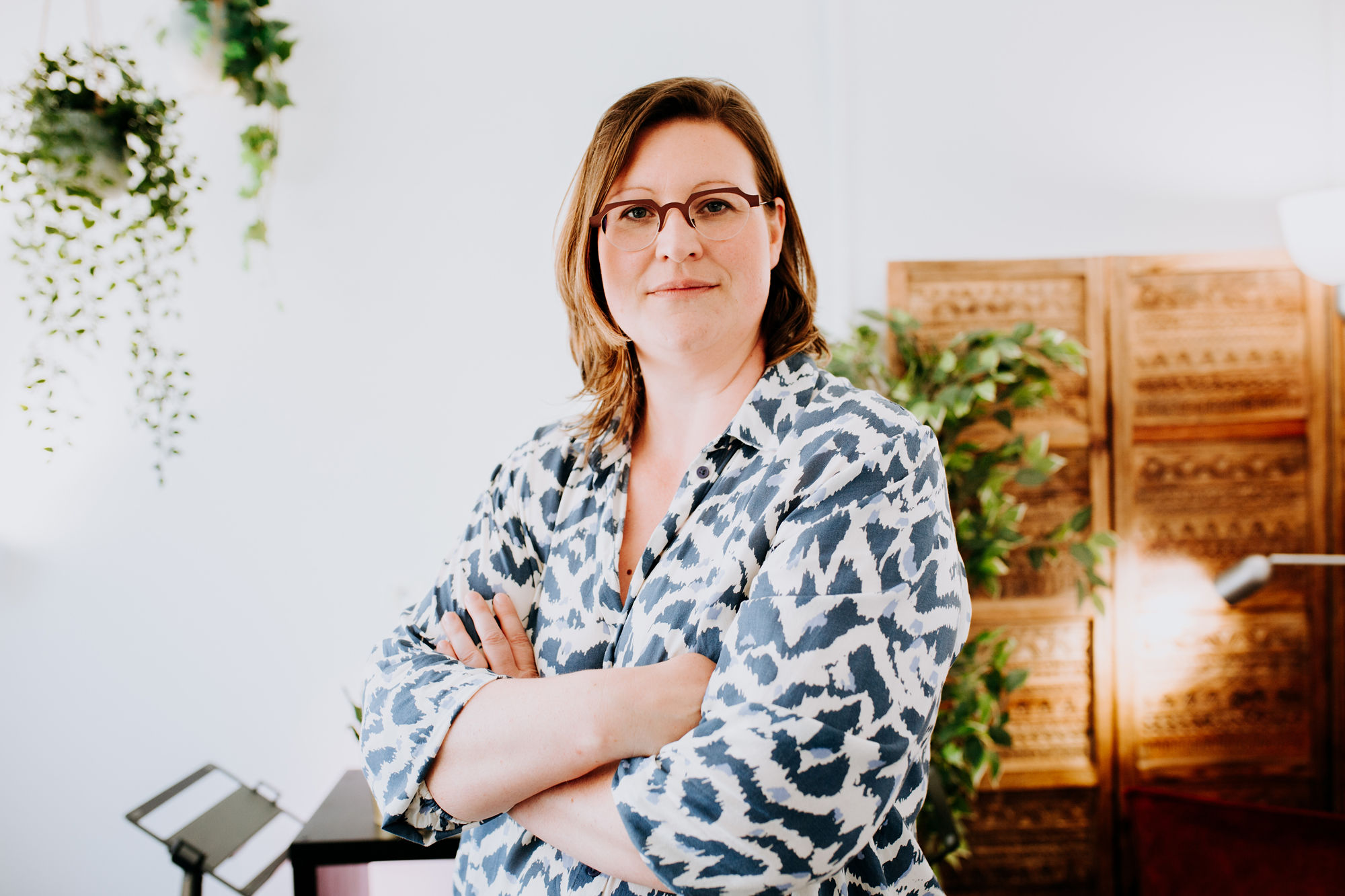 Executive communication coach Ellen Bracquine looking straight at the camera with her arms crossed, a wooden room divider and plants in the background